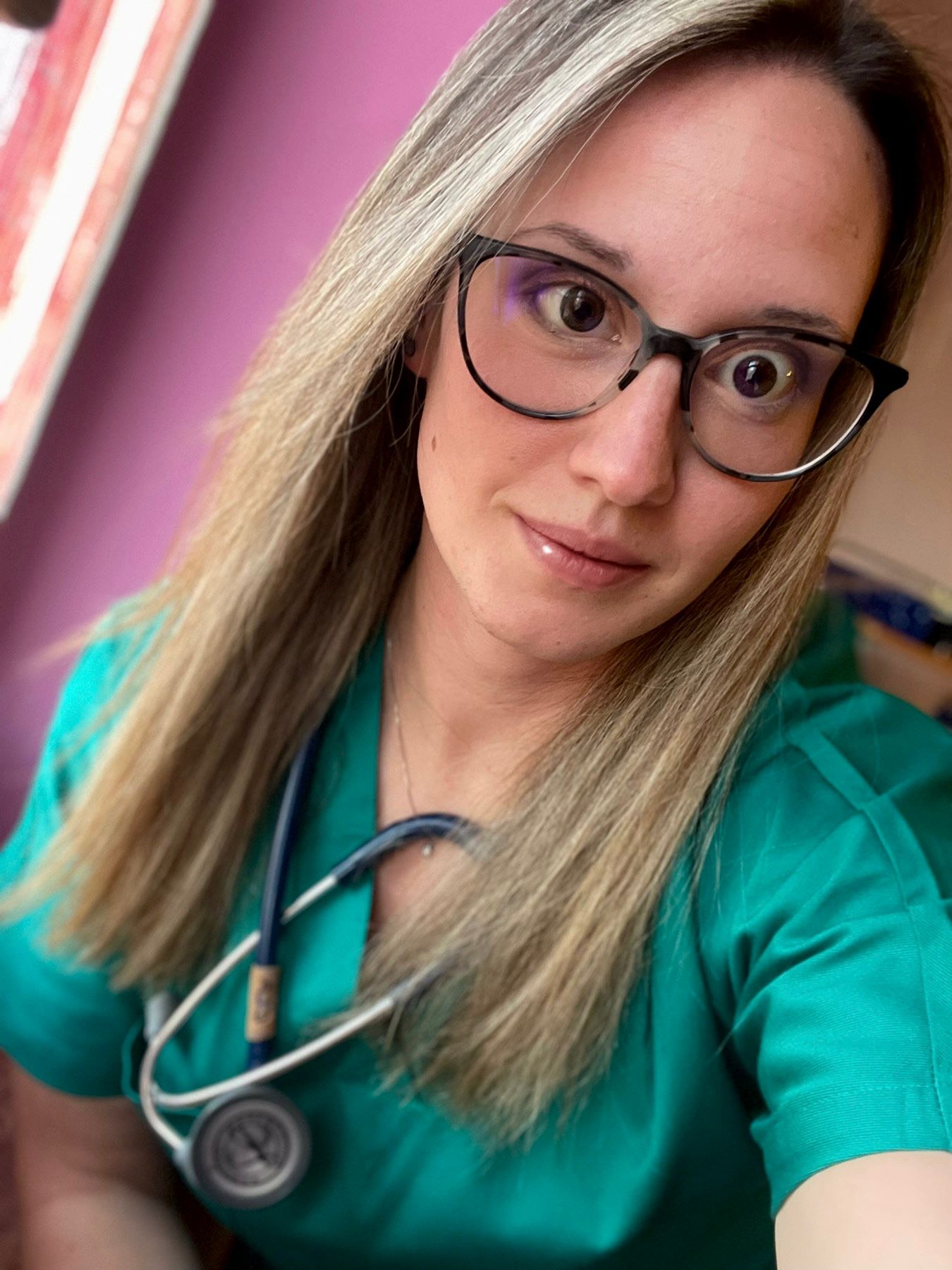 Giulia Bonizzi, con gli occhiali, il camice verde e uno stetoscopio al collo, guarda la telecamera mentre è in piedi davanti a un muro rosa.