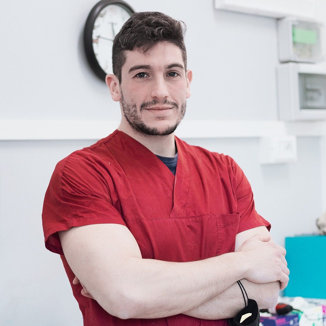 Piero Biassoli è in piedi con le braccia incrociate in un ambiente medico, con indosso un camice rosso. Una mascherina nera gli pende dal polso, con un orologio e materiale medico visibili sullo sfondo.