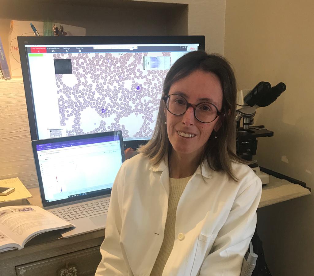 Francesca Massazza, che indossa occhiali e un camice bianco, è seduta a una scrivania con un microscopio, un computer portatile, un libro aperto e un monitor che sullo sfondo mostra quelle che sembrano immagini microscopiche di cellule.