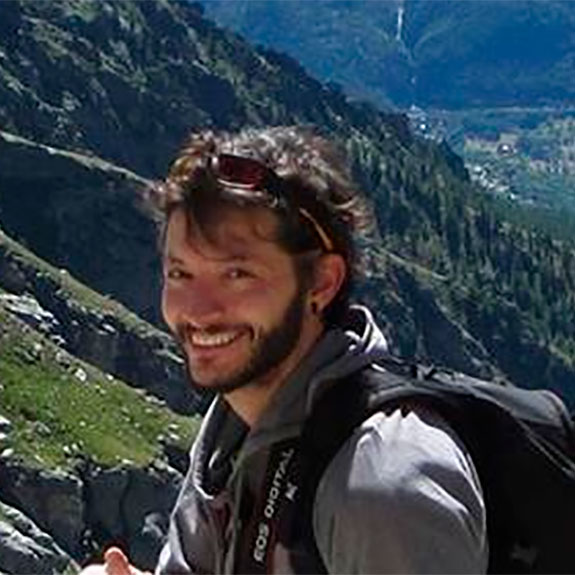 Un uomo con i capelli castani corti e la barba, che indossa una giacca grigia e uno zaino, sorride all'aperto in un paesaggio montuoso con verdi colline e terreno roccioso sullo sfondo, che ricorda lo spirito avventuroso di Giulia Bonizzi.