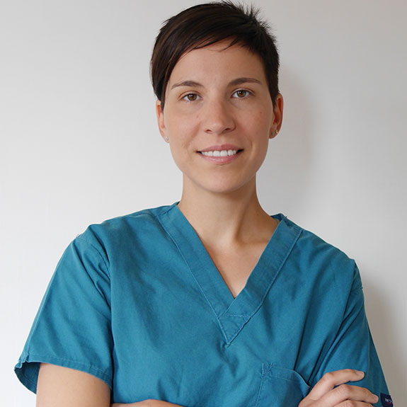 Eleonora Atena Galli, con i capelli corti e scuri, indossa un camice verde acqua ed è in piedi, sorridente e con le braccia incrociate, su uno sfondo bianco.