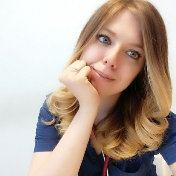 Ilaria Cantamessa, con lunghi capelli biondi e ondulati e occhi azzurri, appoggia il mento sulla mano, sorridendo dolcemente. Indossa una maglietta blu e uno stetoscopio al collo, ed è seduta su uno sfondo bianco.
